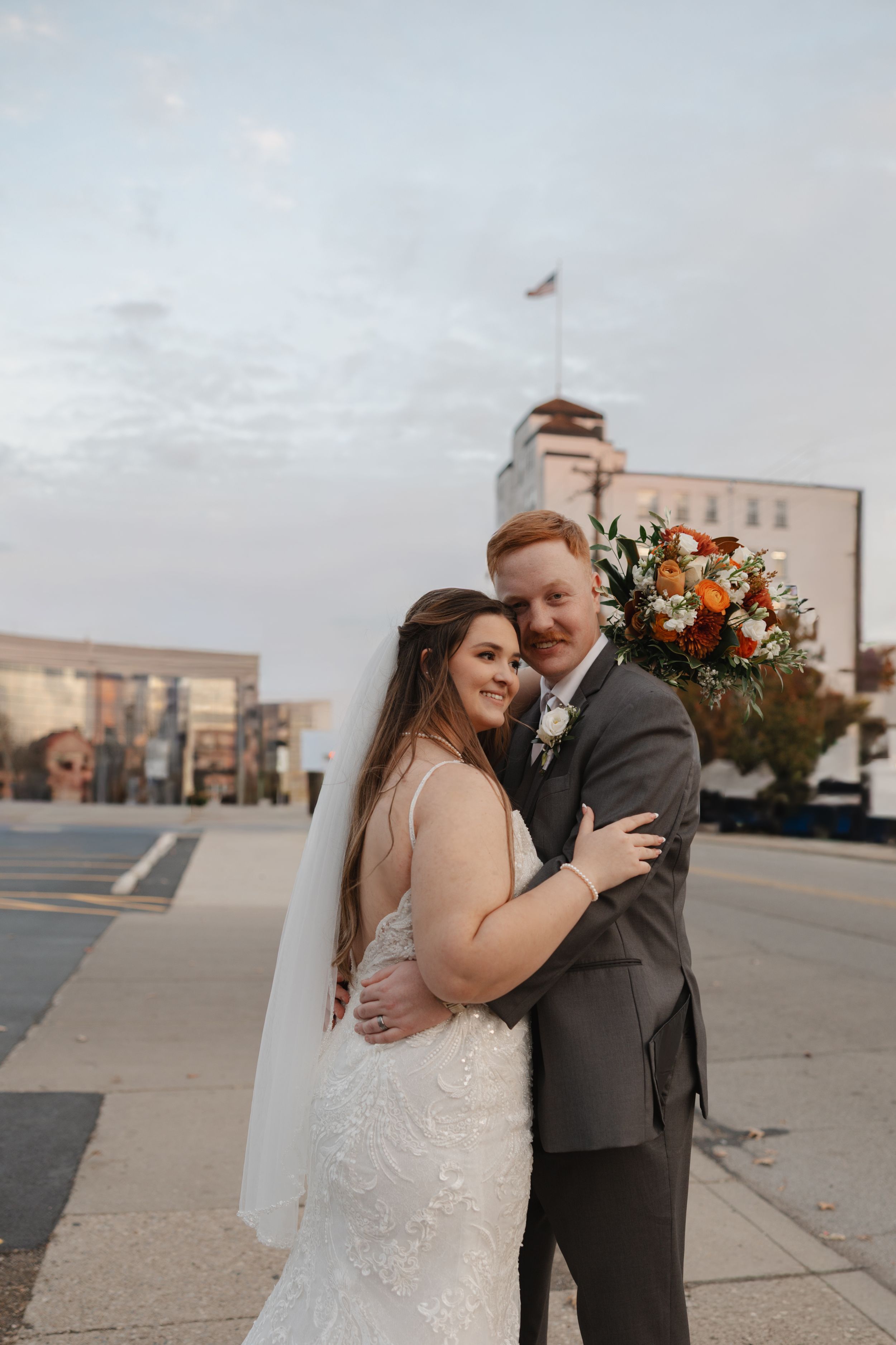 Newlyweds share intimate moments on a rooftop during sunset with urban backdrop and floral bouquet.