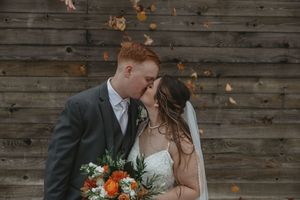 Wedding couple poses against rustic wooden wall backdrop with autumn-toned floral arrangements.