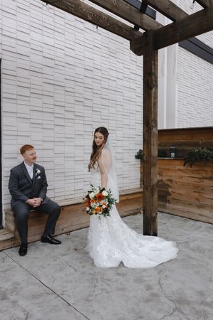 Wedding couple sits and interacts on concrete floor against white brick wall with wooden beams.