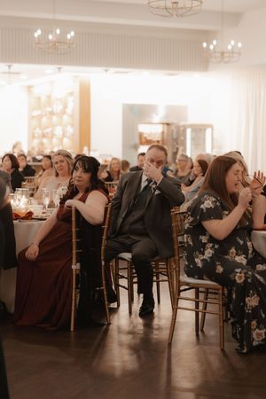 Wedding guests seated at round tables in an elegant reception hall with warm lighting and white decor.