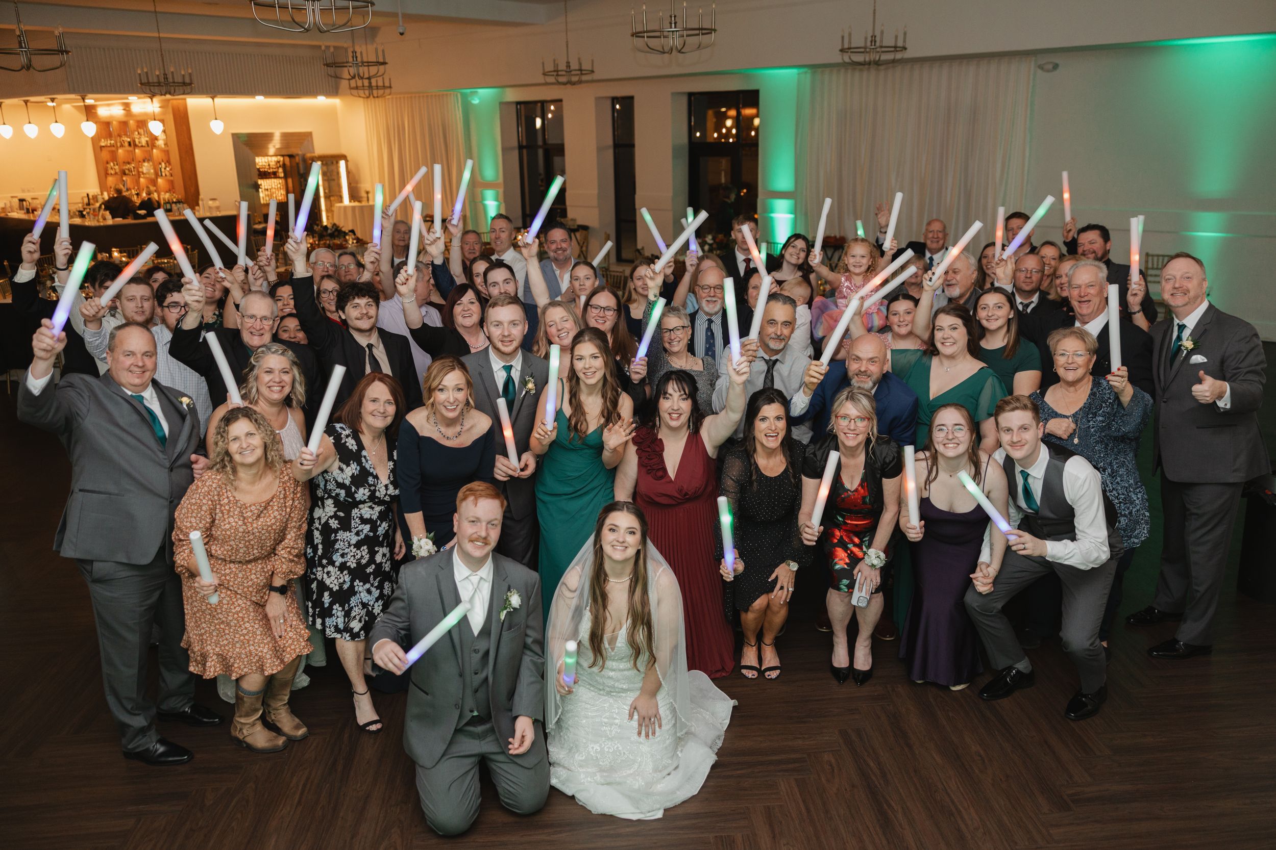 Wedding reception guests dance and celebrate with raised arms in a venue with green lighting.