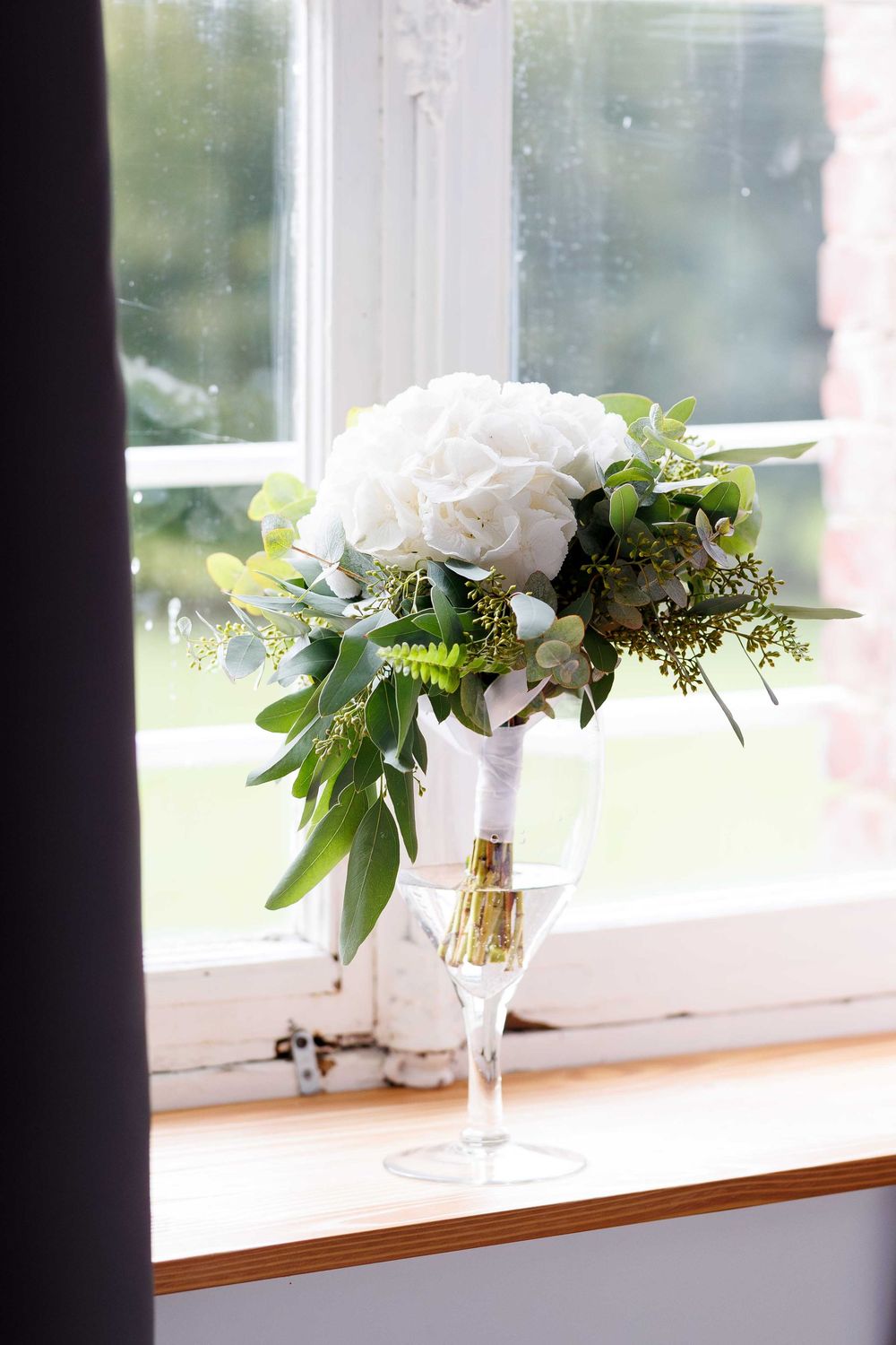 White rose wedding bouquet with greenery accents displayed in a crystal vase on a windowsill.