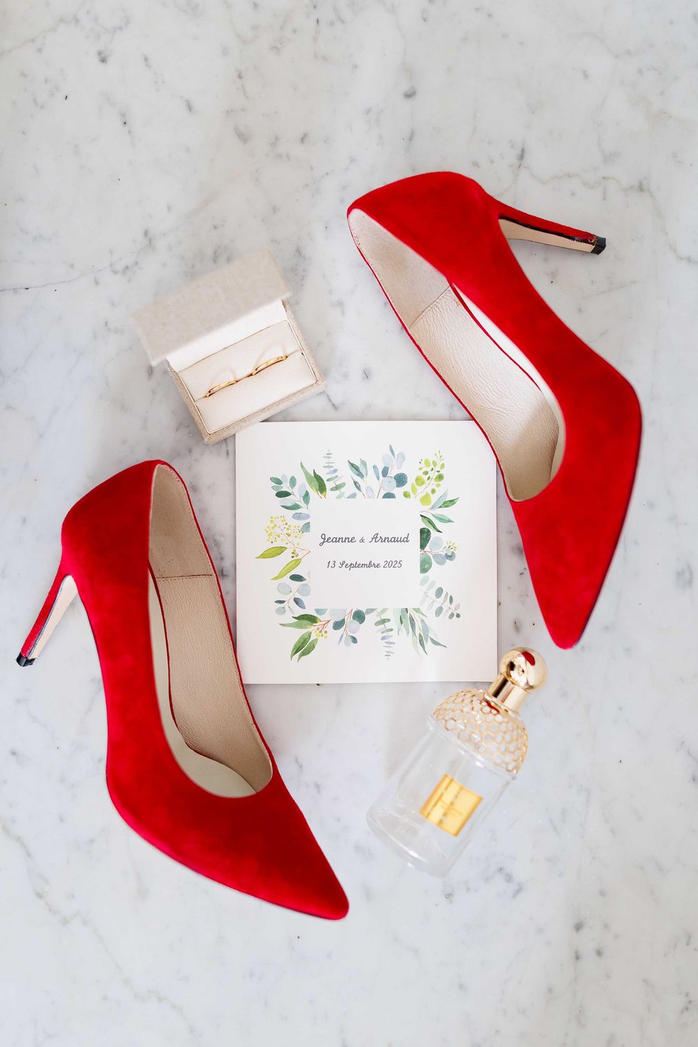 Red stiletto heels and wedding invitation with floral design arranged on marble surface for wedding detail photography.