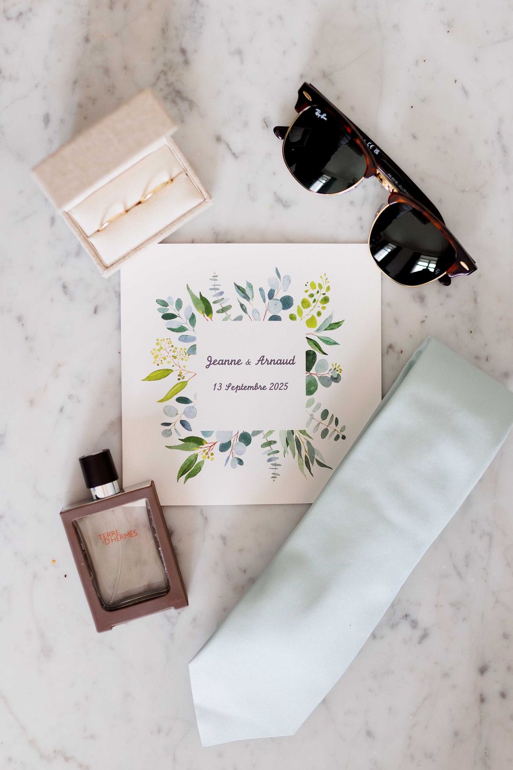 Wedding accessories including invitation with floral design, sunglasses, white tie and perfume bottle on marble surface.