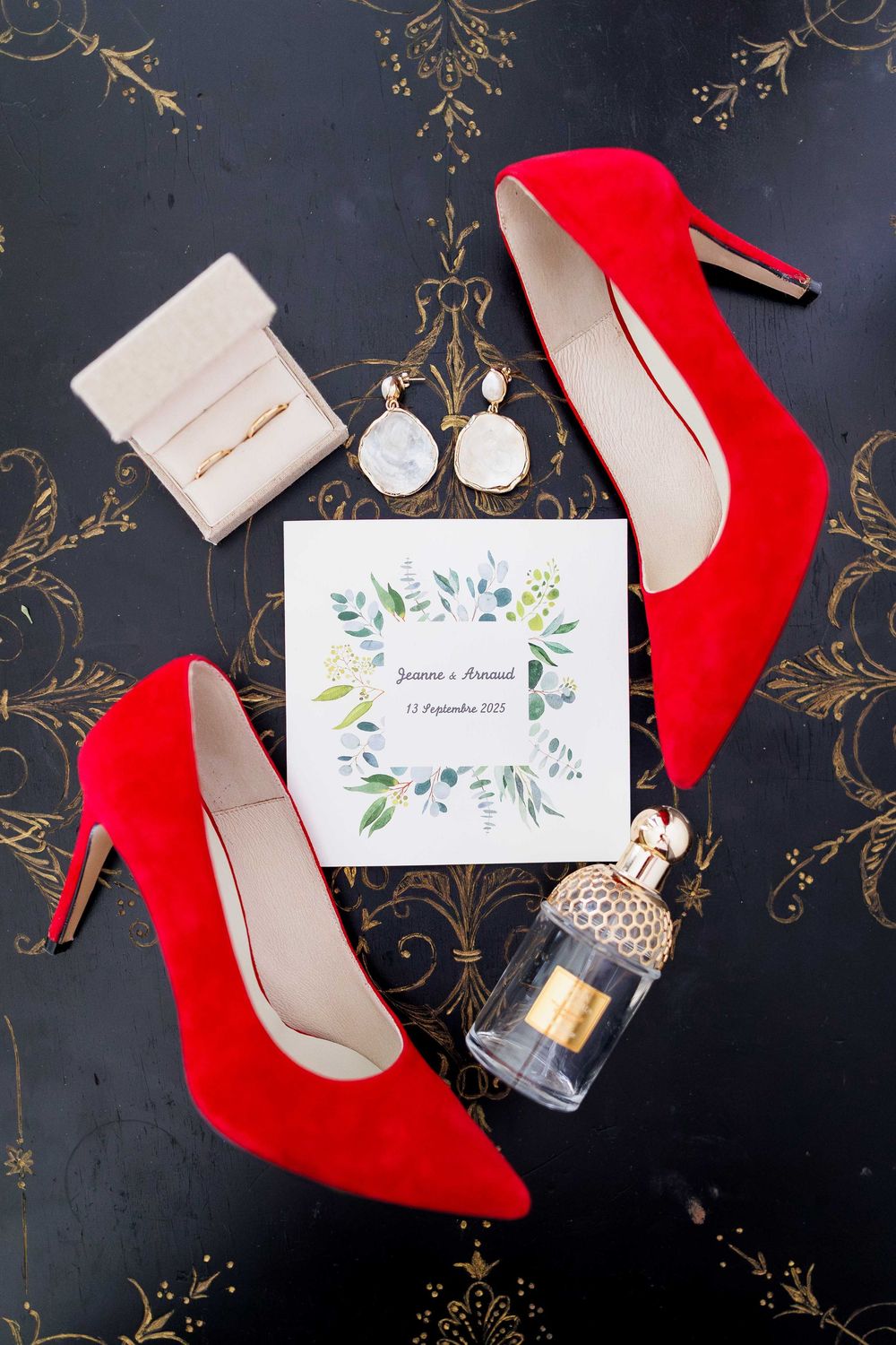 Wedding details flatlay with red stiletto heels, floral invitation, perfume bottle and jewelry on dark background.