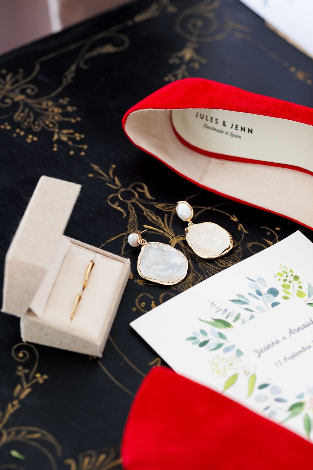 Wedding accessories including red shoes, pearl earrings, and floral invitation card arranged on dark decorative surface.