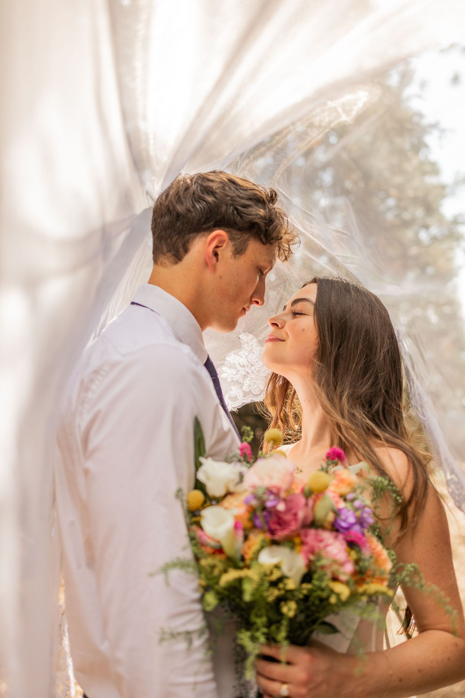 Romantic wedding couple shares intimate kiss under sheer white tent with colorful floral bouquet.