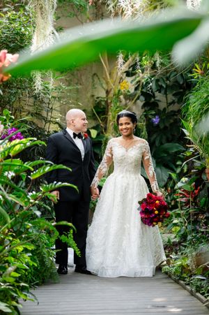 A wedding ceremony in a lush greenhouse setting with white decor and red floral arrangements.