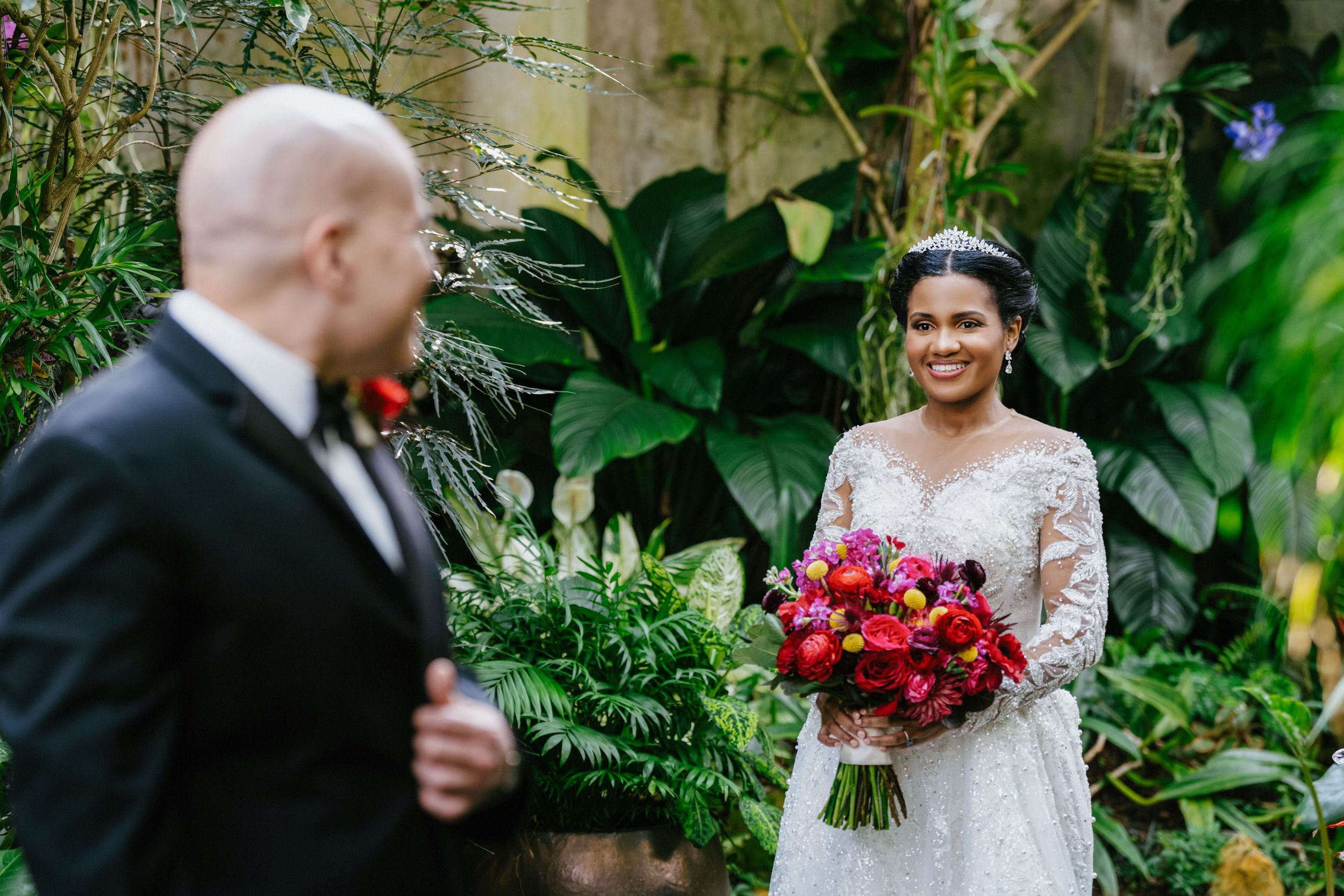 Wedding couple posing among lush greenery with vibrant red floral bouquet.