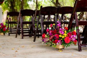Colorful floral arrangements with pink and red roses decorate a rustic outdoor wedding venue with wooden chairs.