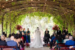 Outdoor wedding ceremony under a natural tree canopy with guests seated on both sides.