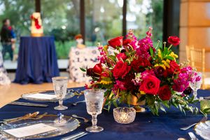 A vibrant red and pink floral centerpiece sits on a navy blue tablecloth with elegant place settings and glassware.