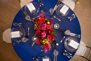 Round navy blue tablecloth with elegant place settings and vibrant red floral centerpieces at a formal event.