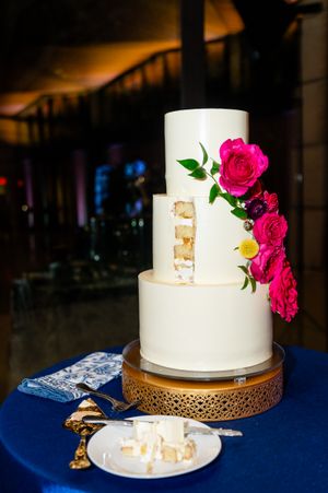 Elegant white wedding cake decorated with vibrant pink roses sits on a gold stand against a navy blue tablecloth.