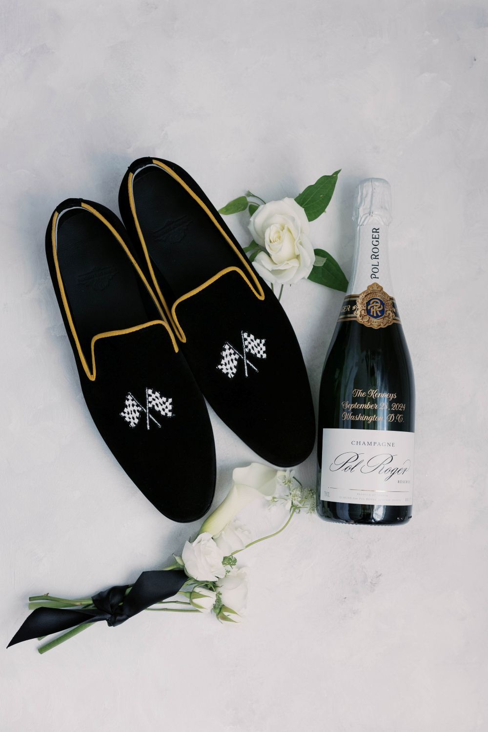 Black velvet slippers with gold embroidery paired with champagne bottle and white roses on marble background.