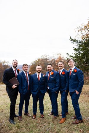 Groomsmen in navy suits and brown shoes pose playfully in a line on a grassy hillside during autumn.