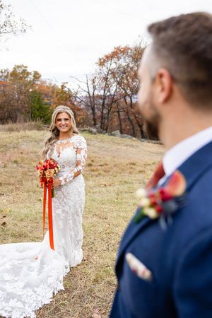 Bride in lace white gown with orange and red bouquet stands in a fall field as groomsmen look on.