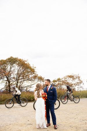 Couple in wedding attire standing on path while cyclists ride by during autumn season. NJ wedding photographer