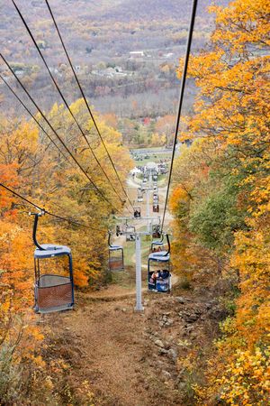 Ski lift chairs glide through vibrant autumn foliage with golden leaves against a mountain landscape backdrop.