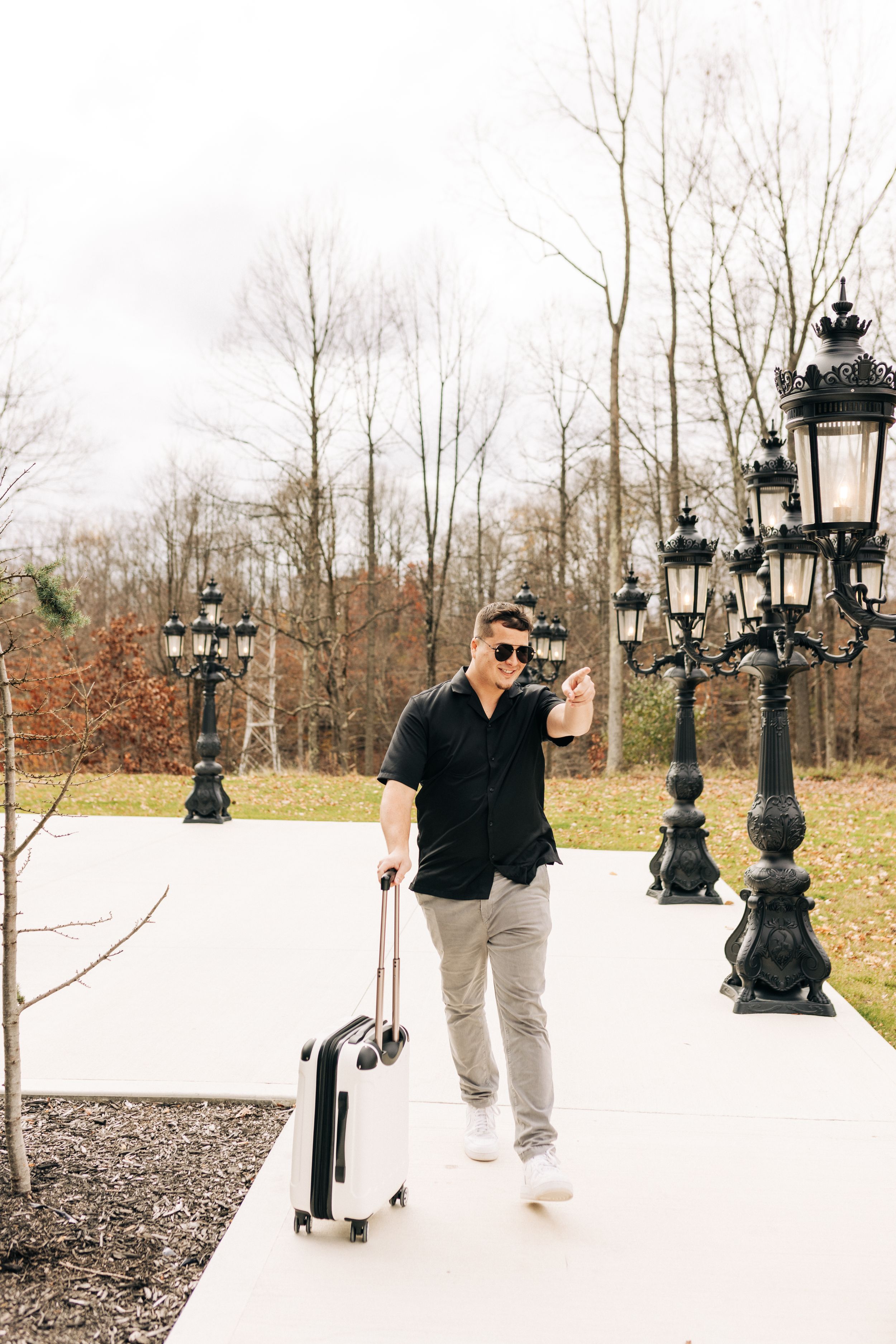 A person in sunglasses and casual attire walks along a white pathway with vintage-style lamp posts and bare trees in background.