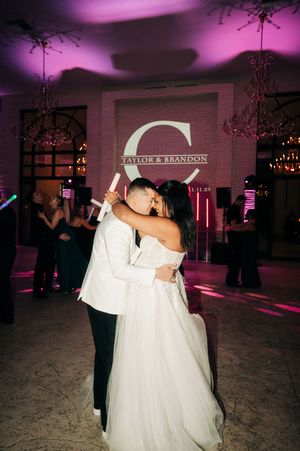 A couple dances together in an elegant ballroom with purple lighting and a chandelier overhead.