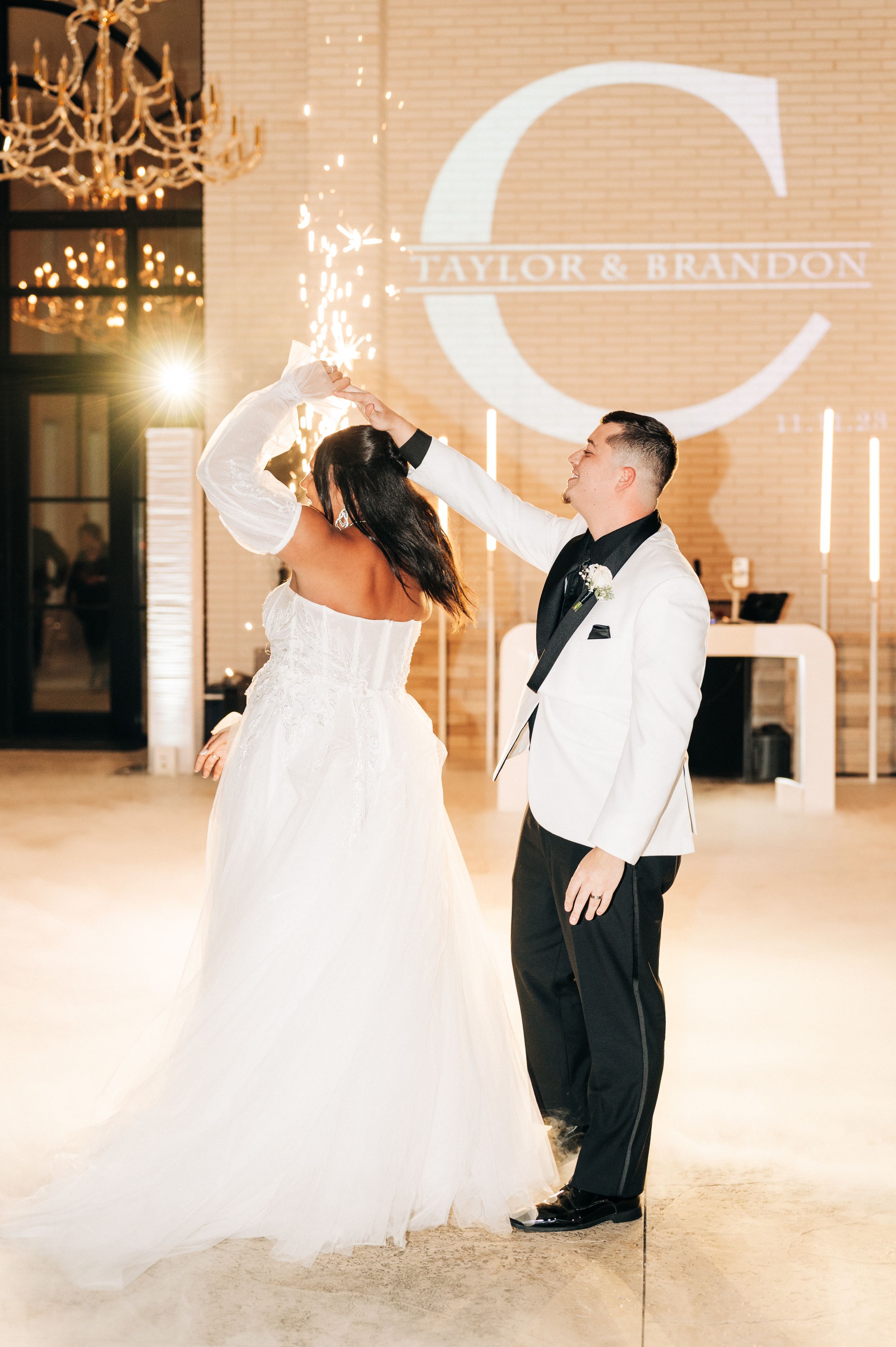 A romantic wedding couple dances together in an elegant ballroom with warm lighting and chandeliers overhead.