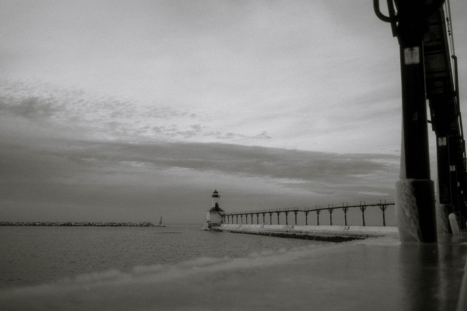 Black and white photograph of a lighthouse at the end of a long pier extending into calm waters under cloudy skies.