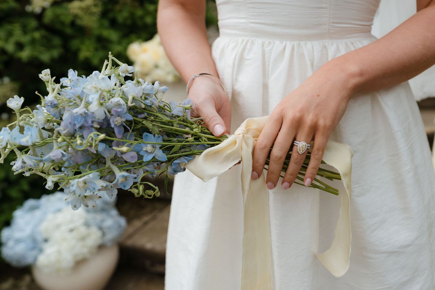 Delicate blue and white wedding bouquet wrapped with cream ribbon held against a white dress.