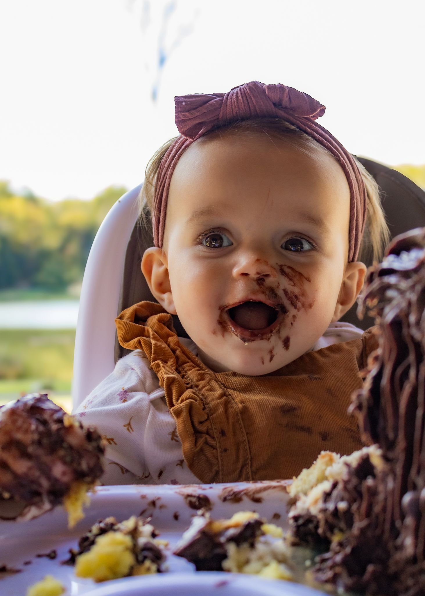 Baby girl with bow headband smiling with chocolate cake smeared on her face during first birthday smash cake.