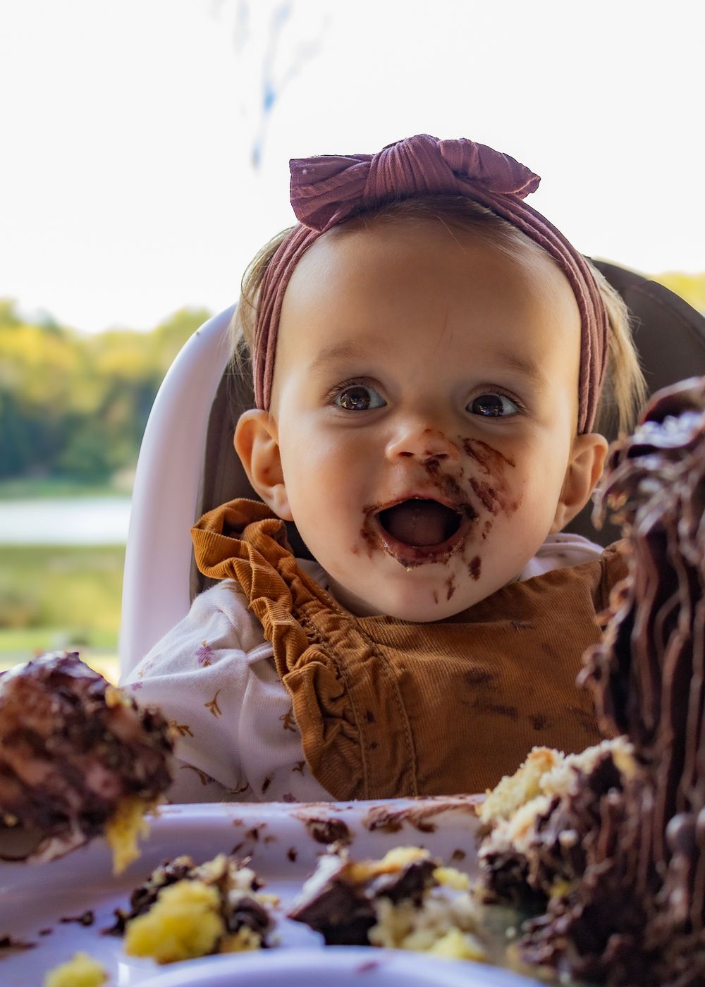 Baby girl with bow headband smiling with chocolate cake smeared on her face during first birthday smash cake.