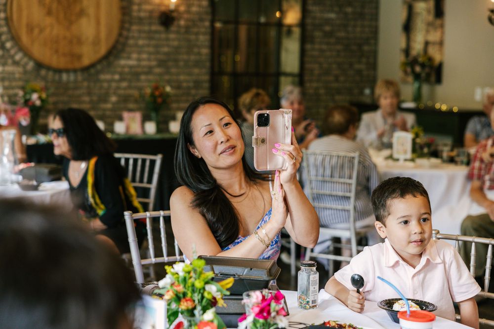 Woman taking photo with pink phone at elegant wedding reception while young boy sits beside her at floral table.