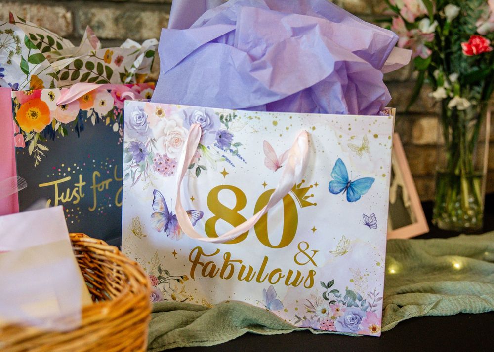 An '80 & Fabulous' gold foil gift bag with butterfly designs surrounded by purple tissue paper and floral decorations.