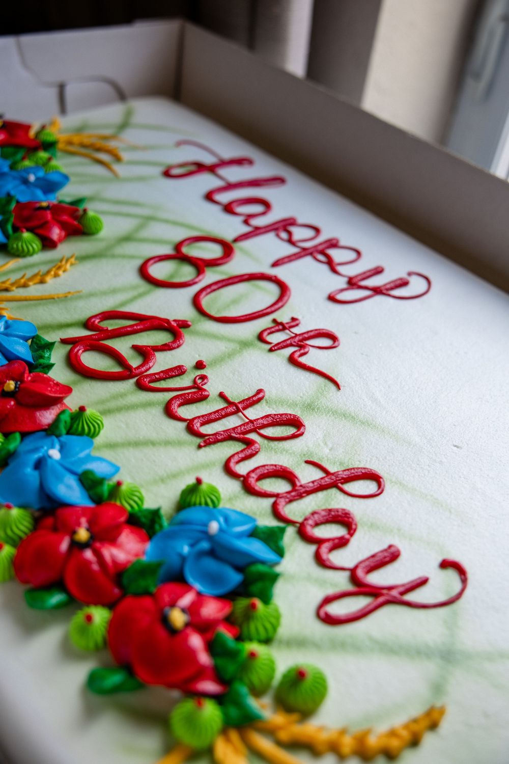 Happy 60th Birthday cake with red script, colorful floral decorations in blue, red, and green frosting on white icing.