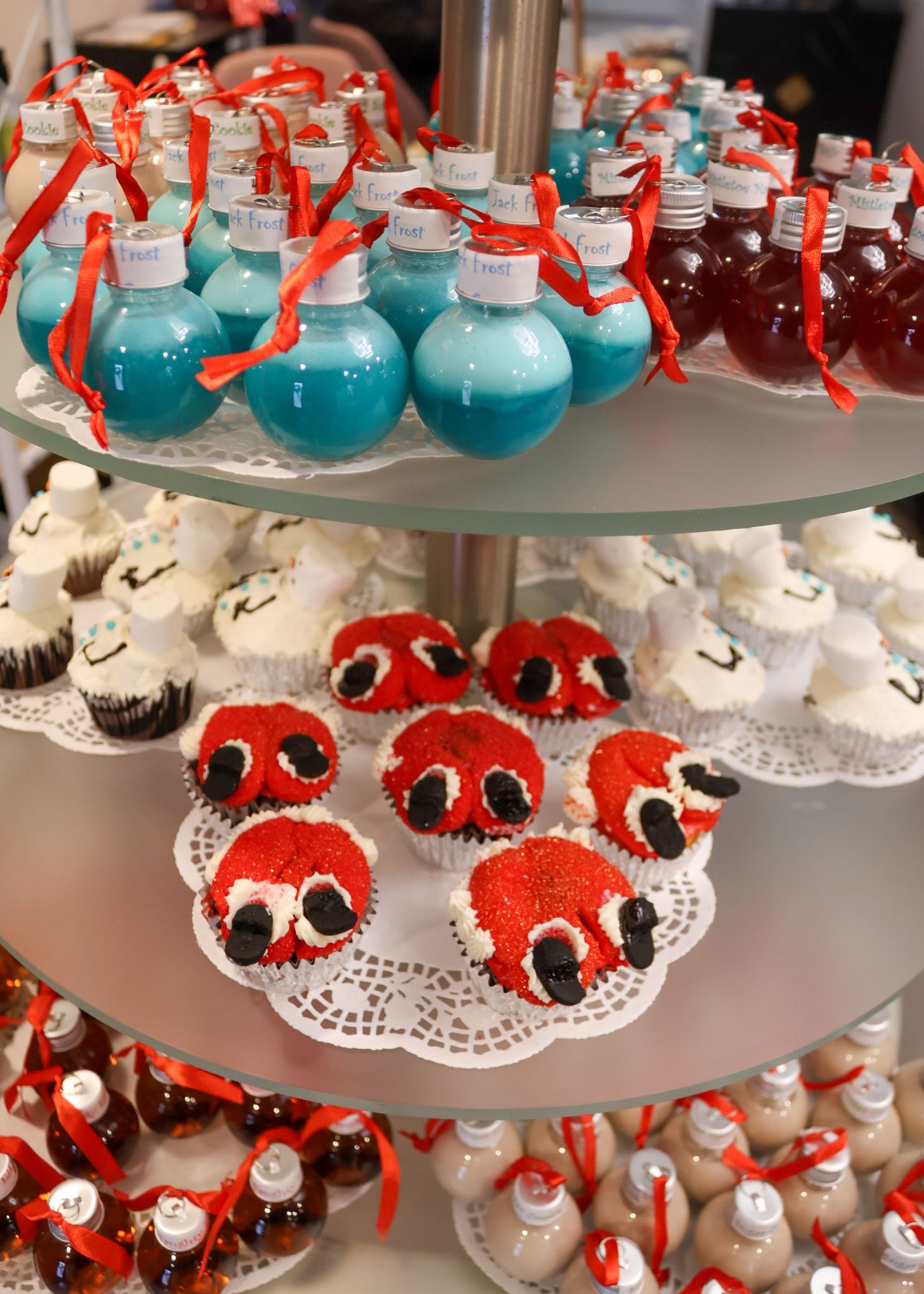 Three-tier dessert stand with red velvet crab cupcakes, blue ornament cake pops, and mini bottles with red ribbons.