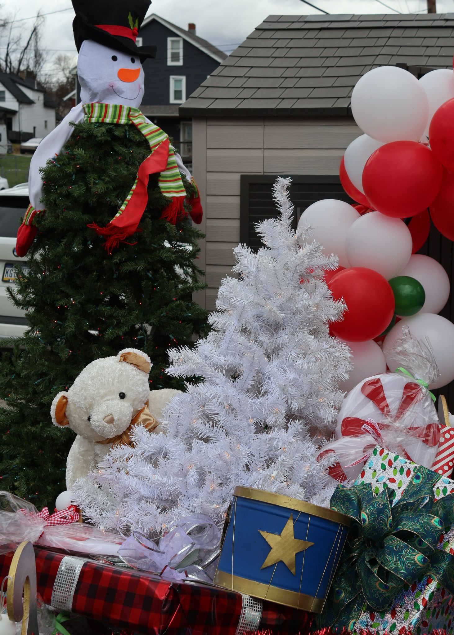 Christmas display with snowman, white tree, teddy bear, balloons, candy canes, and wrapped gifts outdoors.