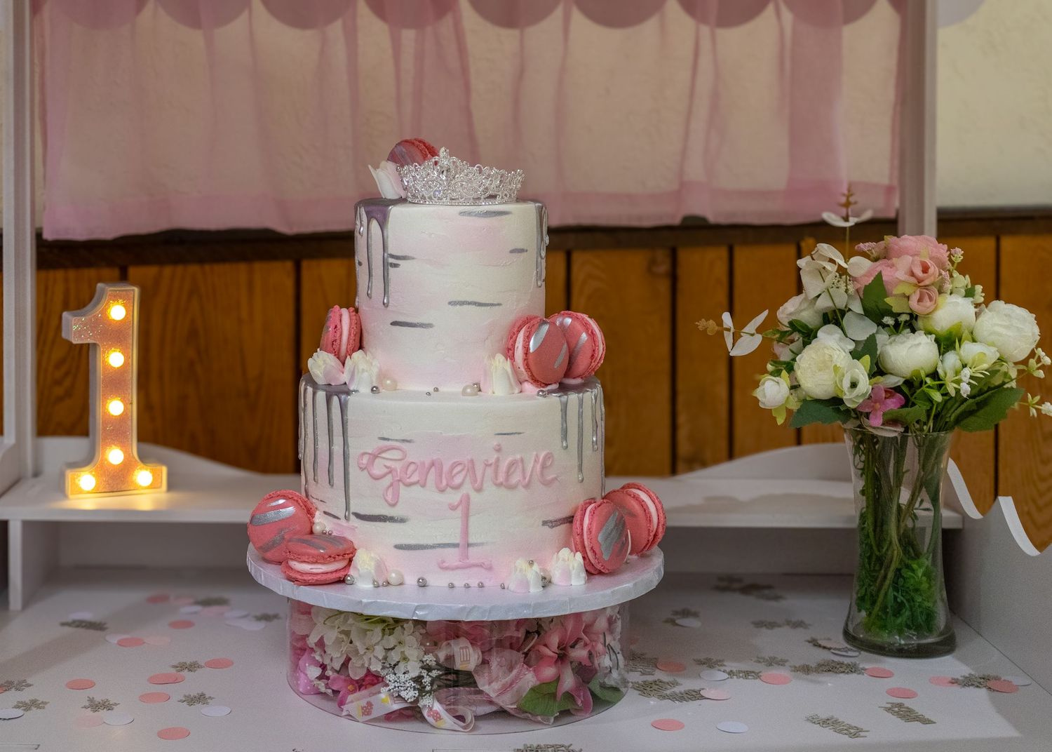 Three-tier white and pink first birthday cake for Genevieve with macarons, crown topper, and floral arrangement.