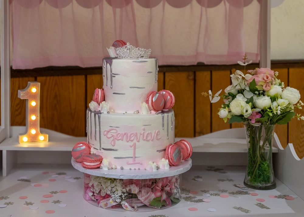 Three-tier white and pink first birthday cake for Genevieve with macarons, crown topper, and floral arrangement.