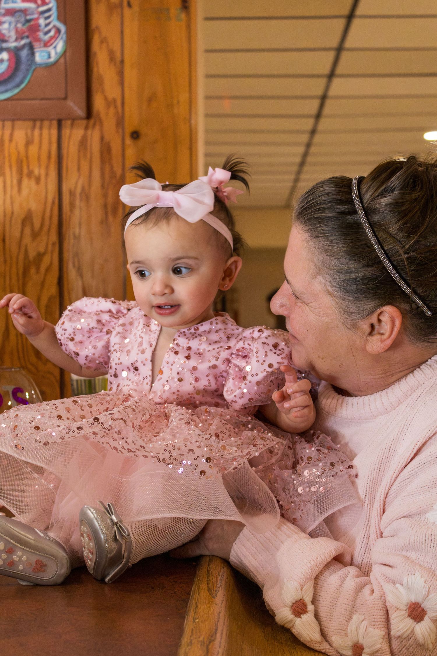Grandmother smiling at baby girl in pink sequin tutu dress with bow headband sitting on wooden bench.