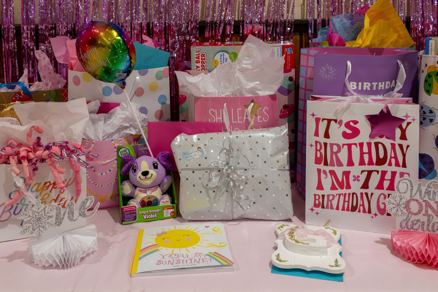 Birthday gifts and bags displayed on a pink table with a colorful foil backdrop and birthday decorations.