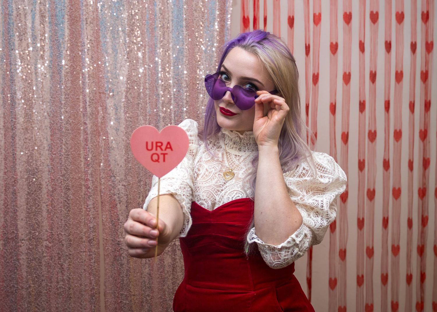 Young woman with purple hair and heart sunglasses holds a pink heart sign reading URA QT at a Valentine's Day photo booth.