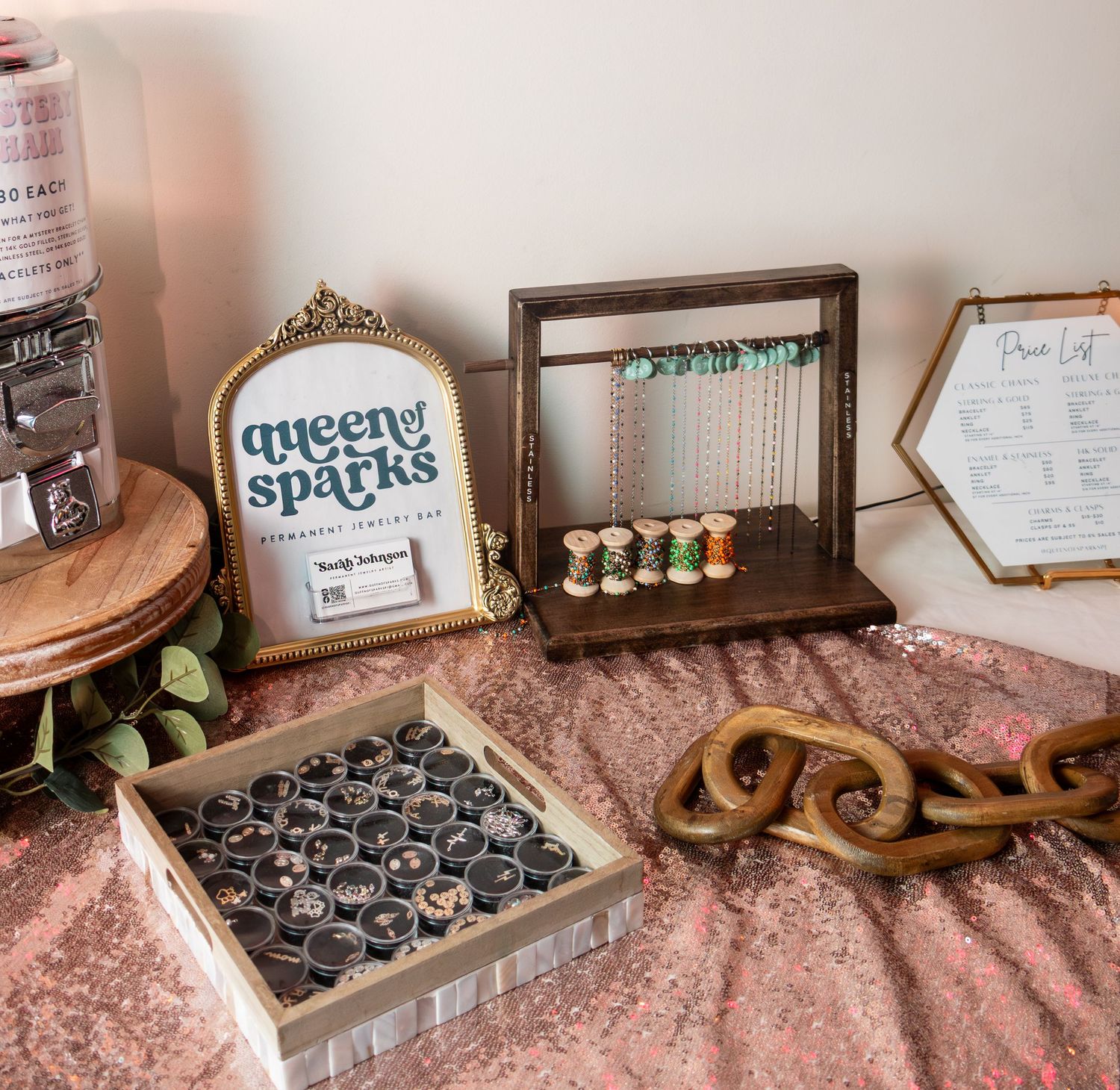 Queen of Sparks jewelry display with rings, necklaces, and price list on a rose gold sequin tablecloth.