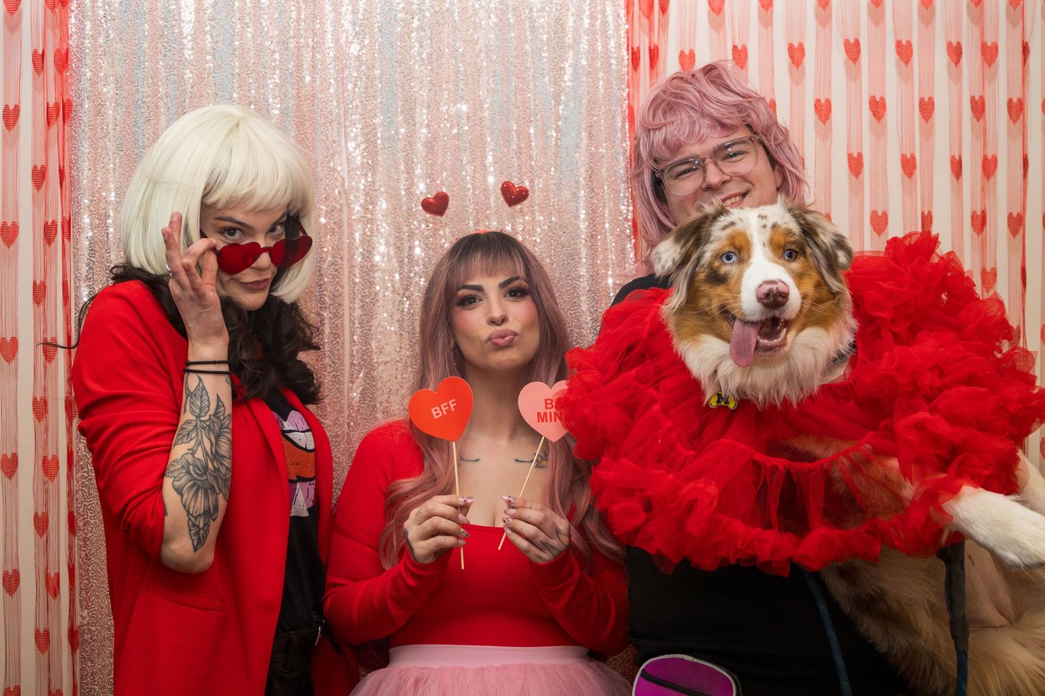 Three people pose in a Valentine's Day photo booth with a dog wearing a red feather boa.