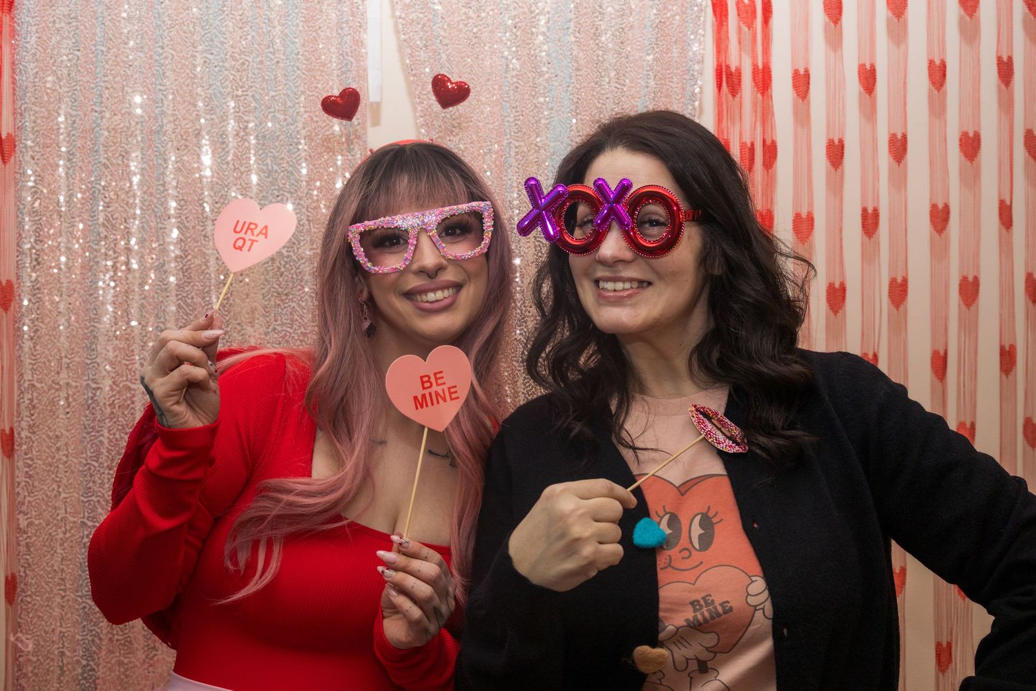 Two women posing in a Valentine's Day photo booth with festive props including XOXO glasses and heart signs.