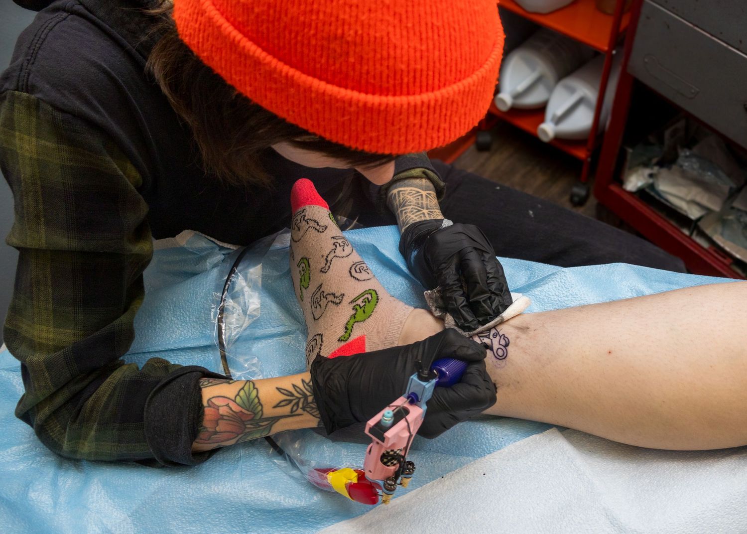 Tattoo artist in orange beanie and black gloves carefully tattooing a client's leg in a studio setting.