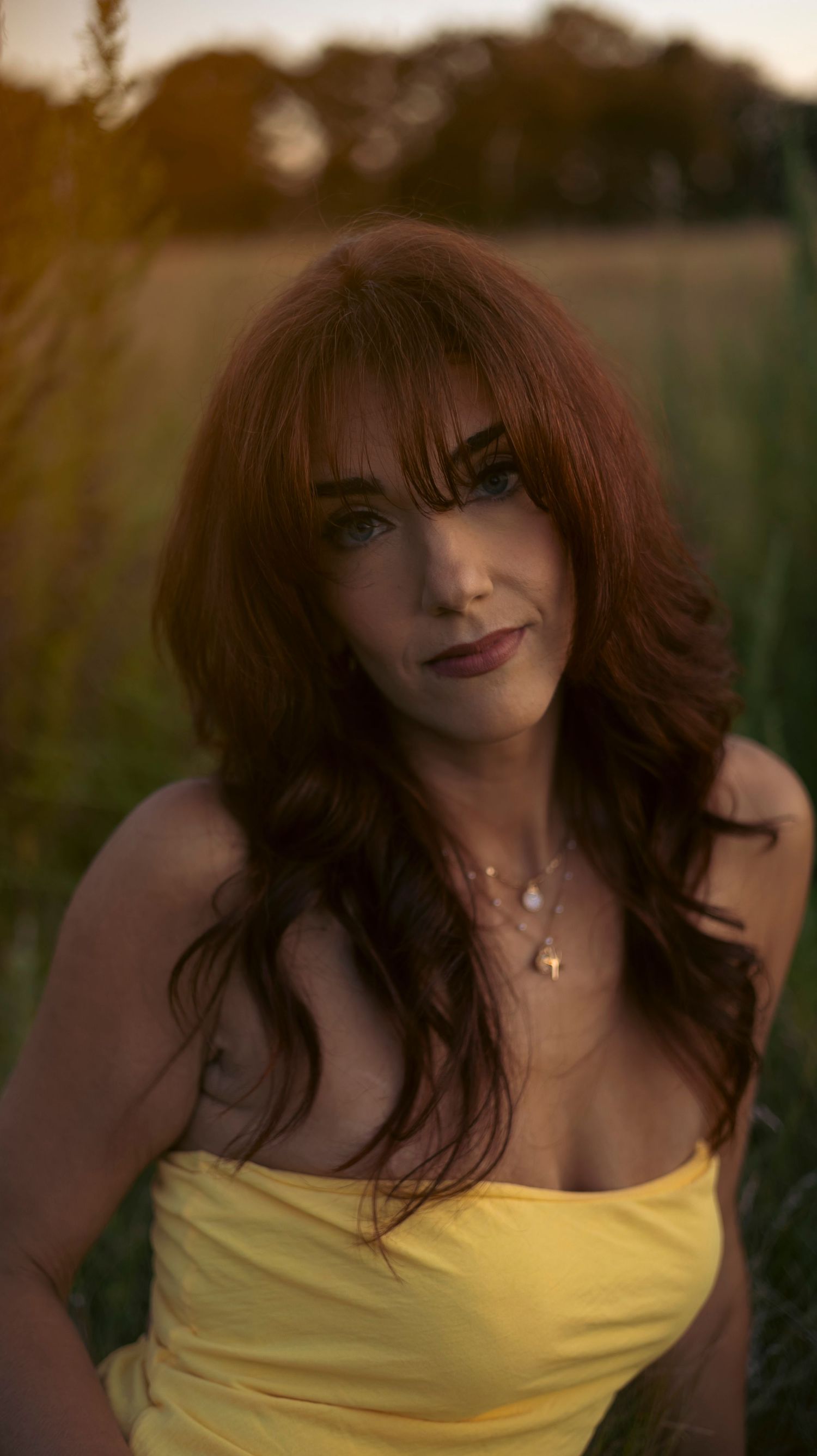 A person with long auburn hair wearing a yellow top poses outdoors during golden hour.