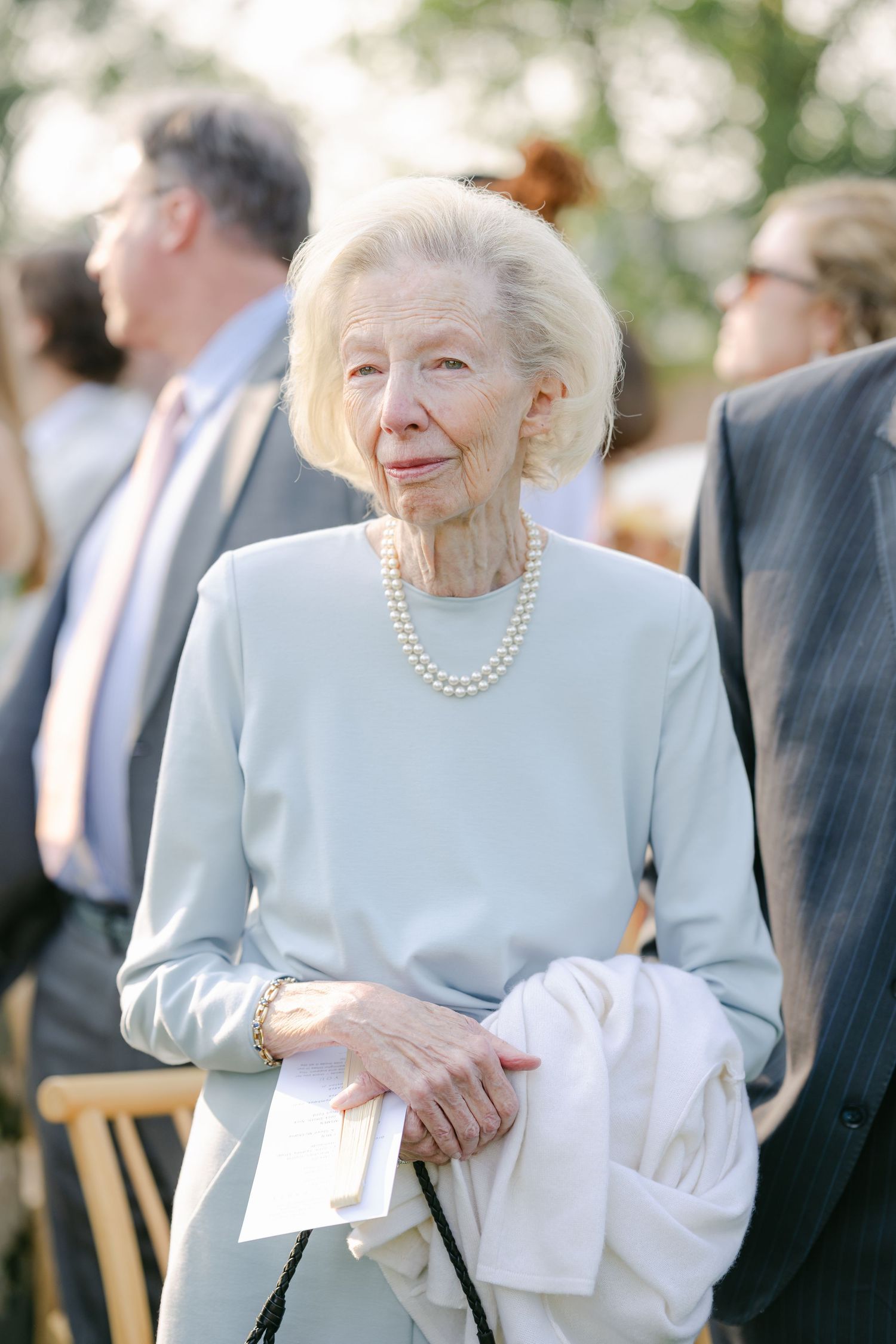An elderly person in a light blue outfit and pearl necklace smiles while attending an outdoor event. - Elawa Farm