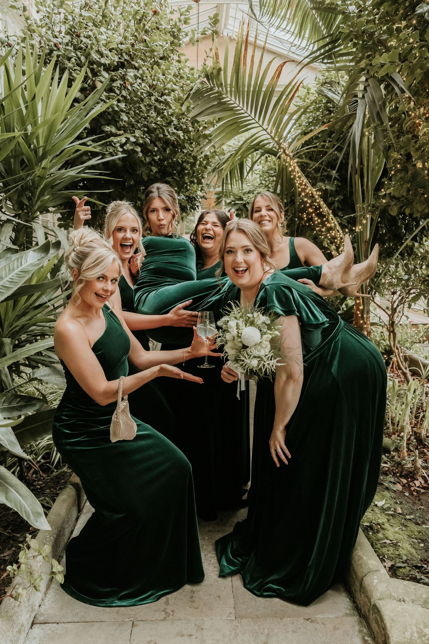 A group in emerald green formal dresses shares candid laughing moments in a lush tropical garden setting.