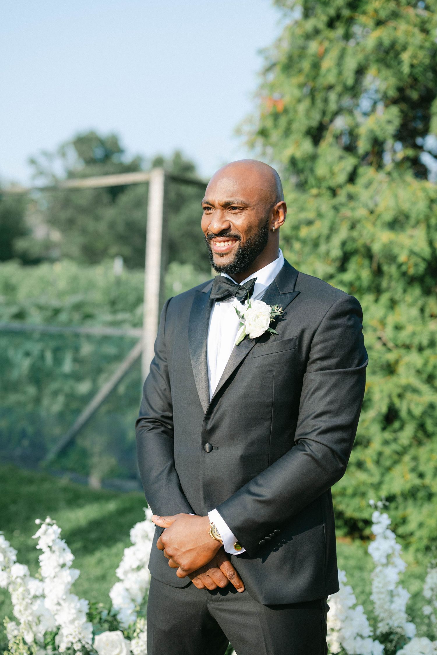 A groom in a classic black tuxedo and bowtie smiles outdoors with white flowers and greenery in the background. - Elawa Farm