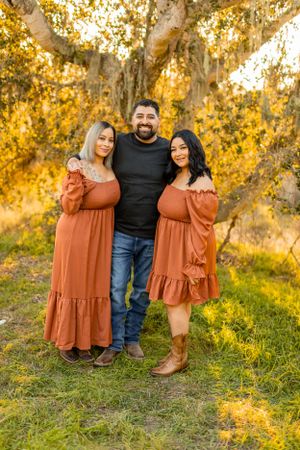 Three people in coordinating rust-colored dresses pose together in autumn setting.