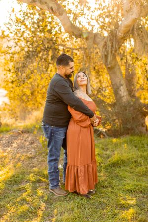 Couple in autumn attire embraces on grassy hillside during golden hour.
